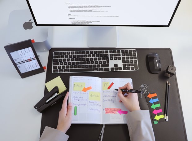 Eine Person plant ihr Praktikum mithilfe eines Terminkalenders. Sie sitzt vor einem Computer und ist von Bürobedarf umgeben.