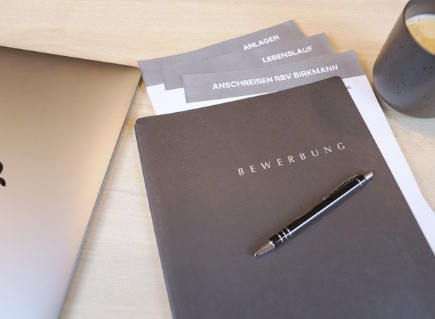 Auf einem Tisch liegt eine Bewerbungsmappe, aus der verschiedene Dokumente herausragen, auf ihr liegt ein Kugelschreiber.