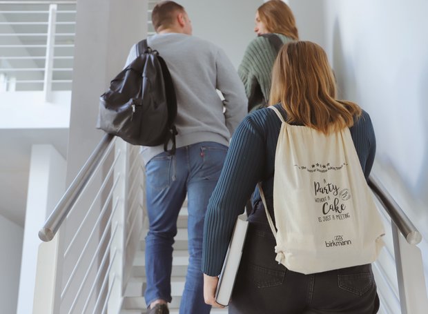 Studierende gehen eine Treppe hinauf. Eine Person trägt ein Buch unter dem Arm und hat einen Rucksack von RBV Birkmann auf.