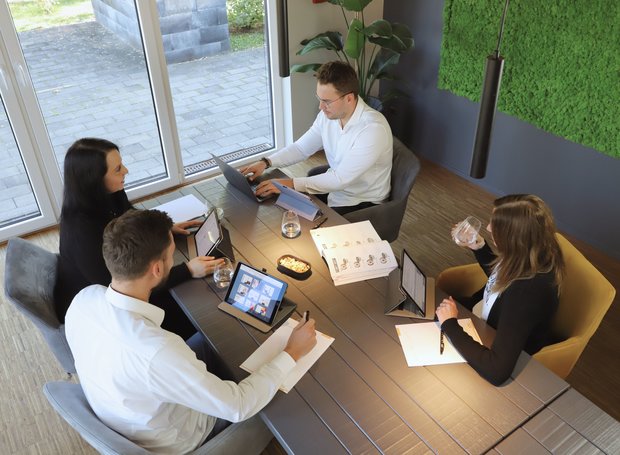 Vier Personen befinden sich in einem Business Meeting und besprechen etwas. Sie haben Tablets, Laptops und Kataloge um sich.